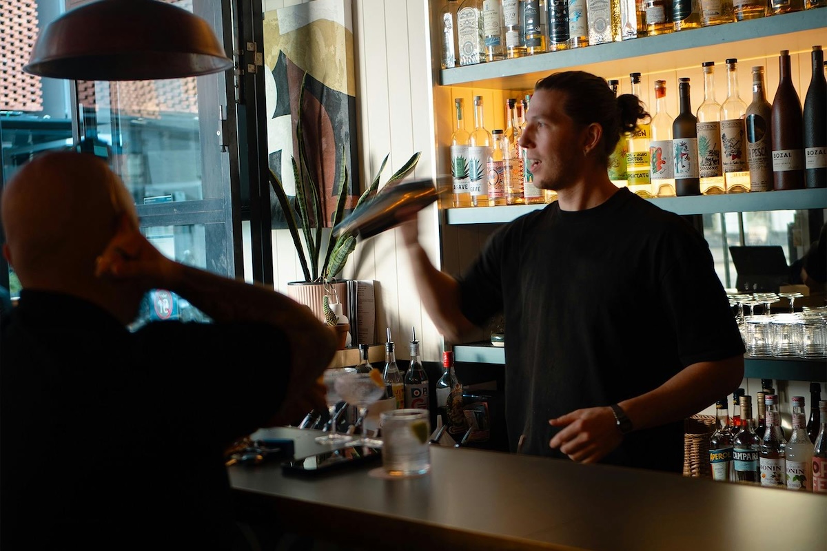 Bartender shaking a cocktail behind a fully stocked bar counter at Bar Bar as a customer watches.
